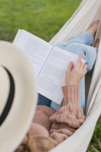 Woman in a hammock wearing a beige sweater and hat, enjoying a book outdoors.